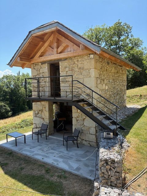 Maisonnette en pierre avec toit en bois, escalier extérieur et petite terrasse avec chaises dans un jardin gazonné