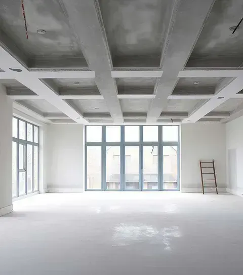 Habitación moderna vacía con vigas blancas en el techo, grandes ventanales y una escalera apoyada contra la pared.