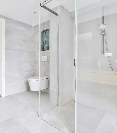 Baño moderno y luminoso con ducha de cristal, azulejos blancos, inodoro suspendido y cabezal de ducha tipo lluvia.