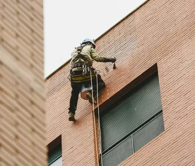 Un operario que trabaja mediante acceso por cuerda desciende por la pared de ladrillo de un edificio junto a una ventana.