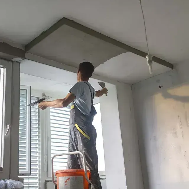 Un trabajador pinta el techo blanco de una habitación, subido a una escalera de mano cerca de una ventana.
