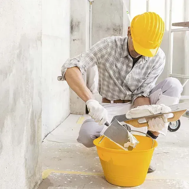 Obrero de la construcción con casco amarillo pintando una pared con un cubo de pintura cerca