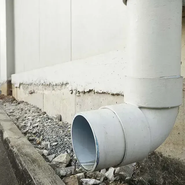Codo de bajante blanco junto a la pared de un edificio sobre un lecho de grava.