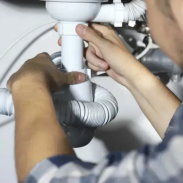 Manos reparando tuberías blancas debajo de un fregadero con una manguera de desagüe flexible gris.