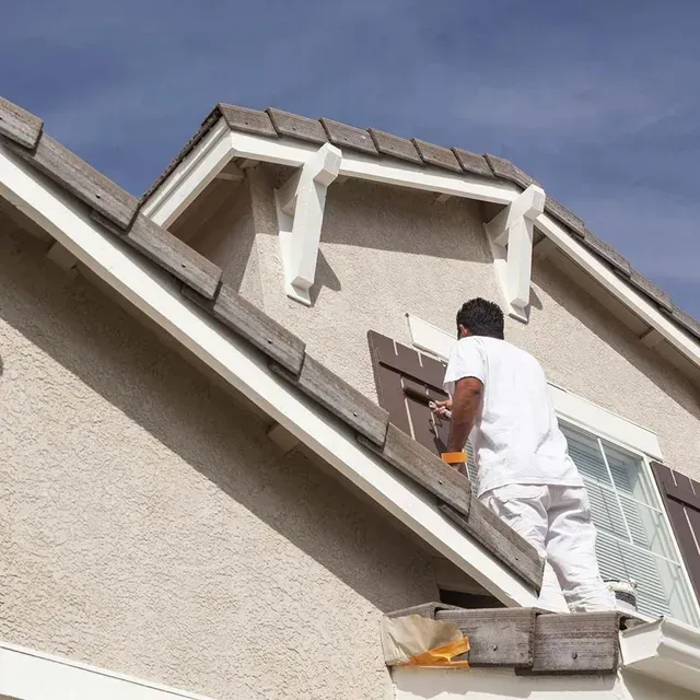 Un trabajador pinta el exterior de una casa mientras está subido a una escalera cerca del tejado.
