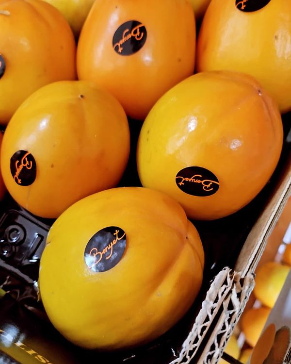Primer plano de caquis maduros de color naranja con etiquetas negras, apilados en un recipiente de cartón.