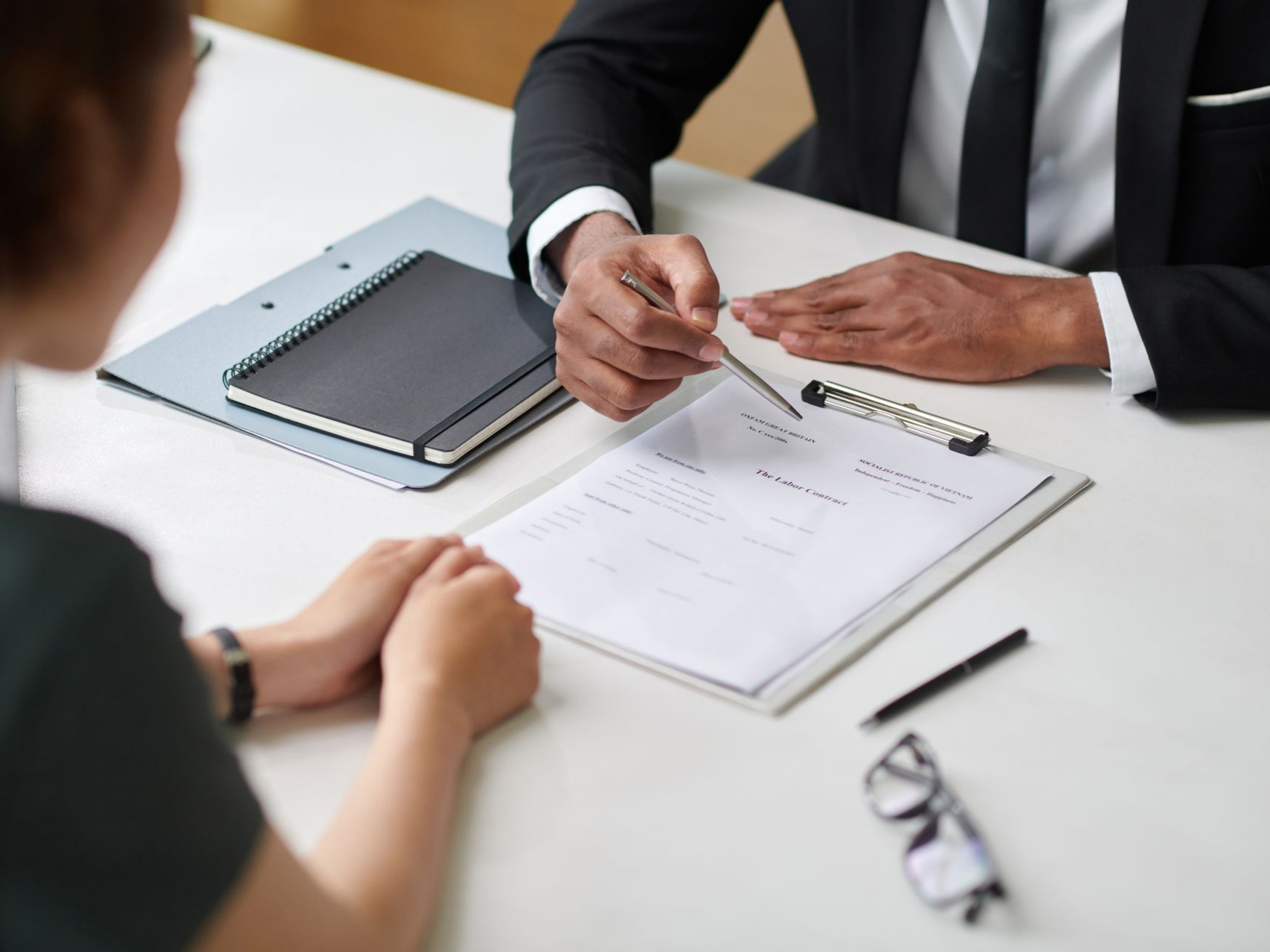 Une personne en costume désigne des documents du doigt avec une autre personne, assise à une table, discute de ces documents.