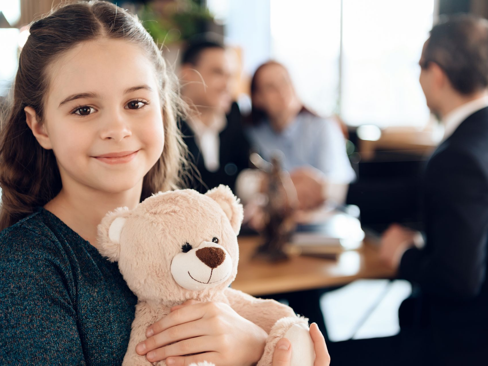 Une fillette sourit, tenant un ours en peluche devant une famille et son avocat.