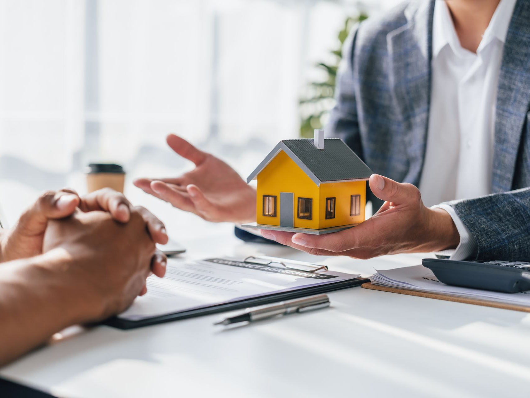 Un agent immobilier présente une petite maison témoin jaune à un client ; des documents et une calculatrice sont posés sur la table.