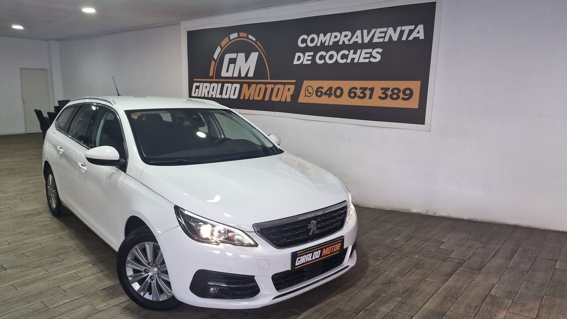 Peugeot 308 SW blanco estacionado en el interior frente a un cartel de "Venta de autos" con un número de teléfono.