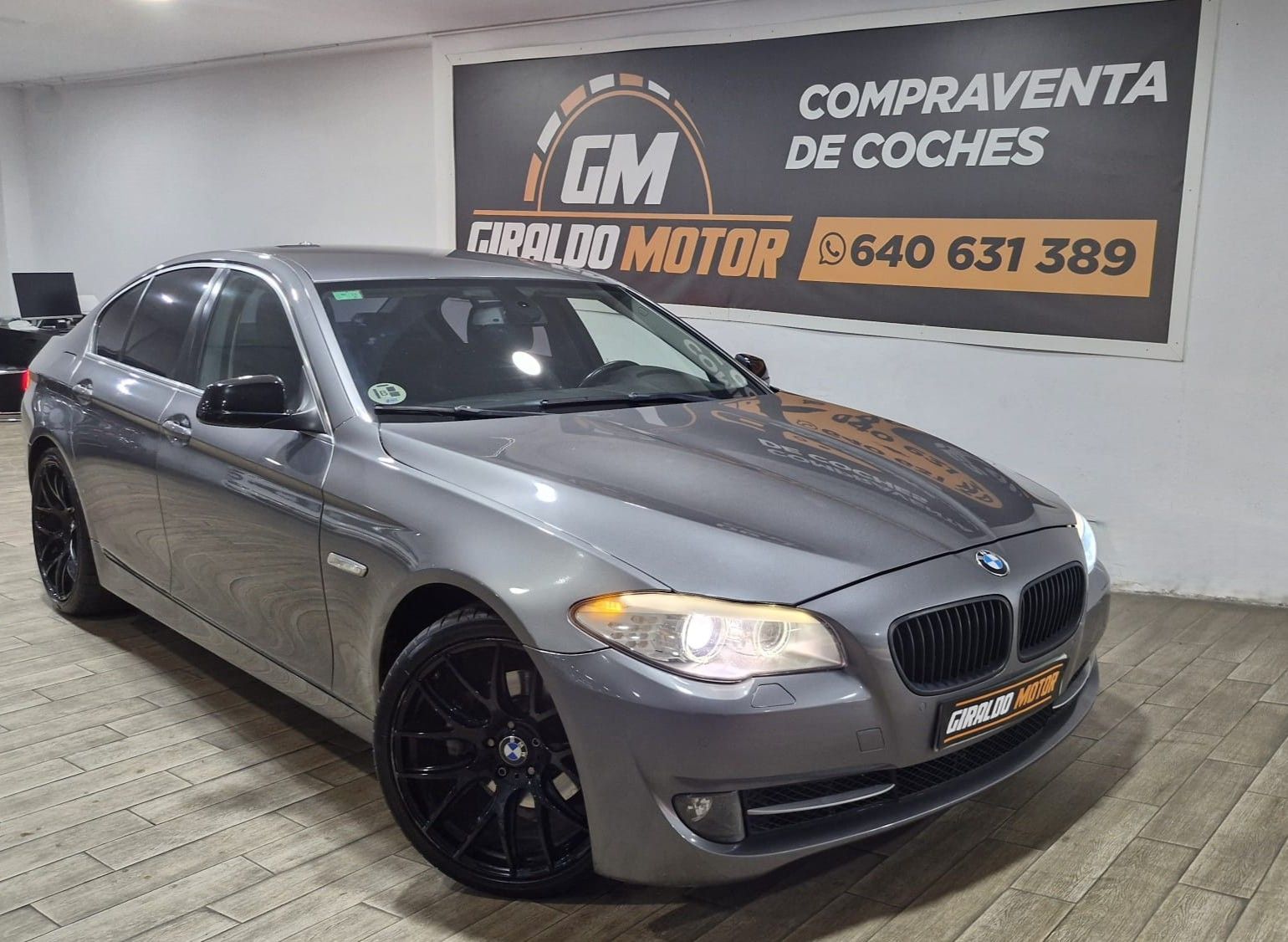 BMW sedán gris estacionado en un concesionario, vista frontal. Llantas negras. Cartel de "Compraventa de coches" detrás.