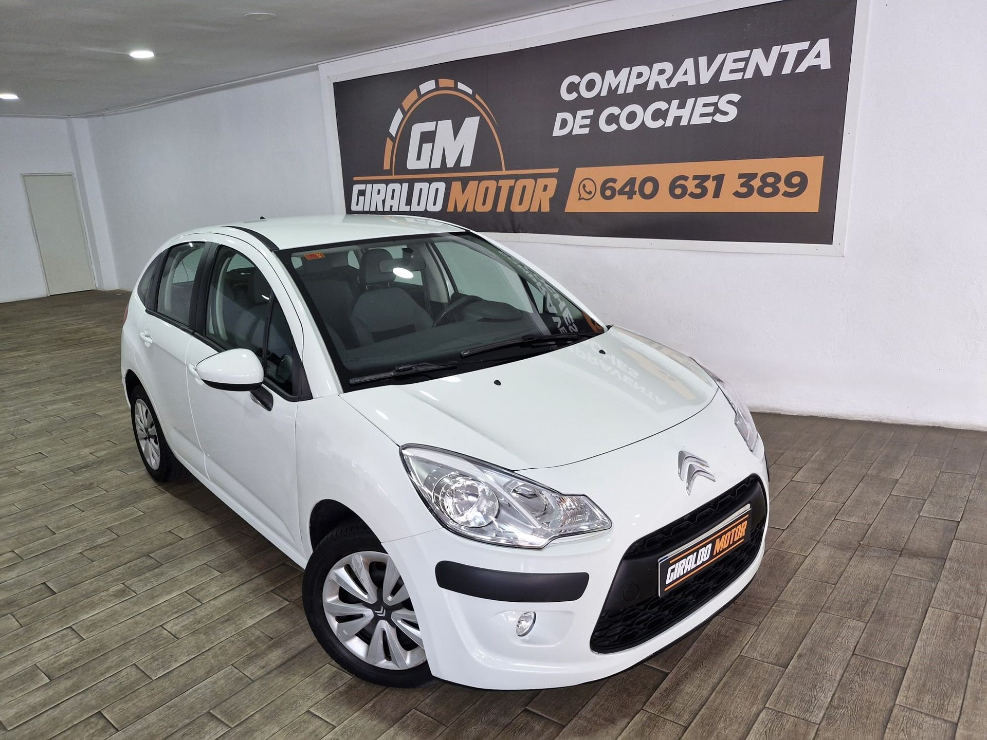 Citroën C3 hatchback blanco estacionado en el interior frente a un cartel de “COMPRAVENTA DE COCHES”.