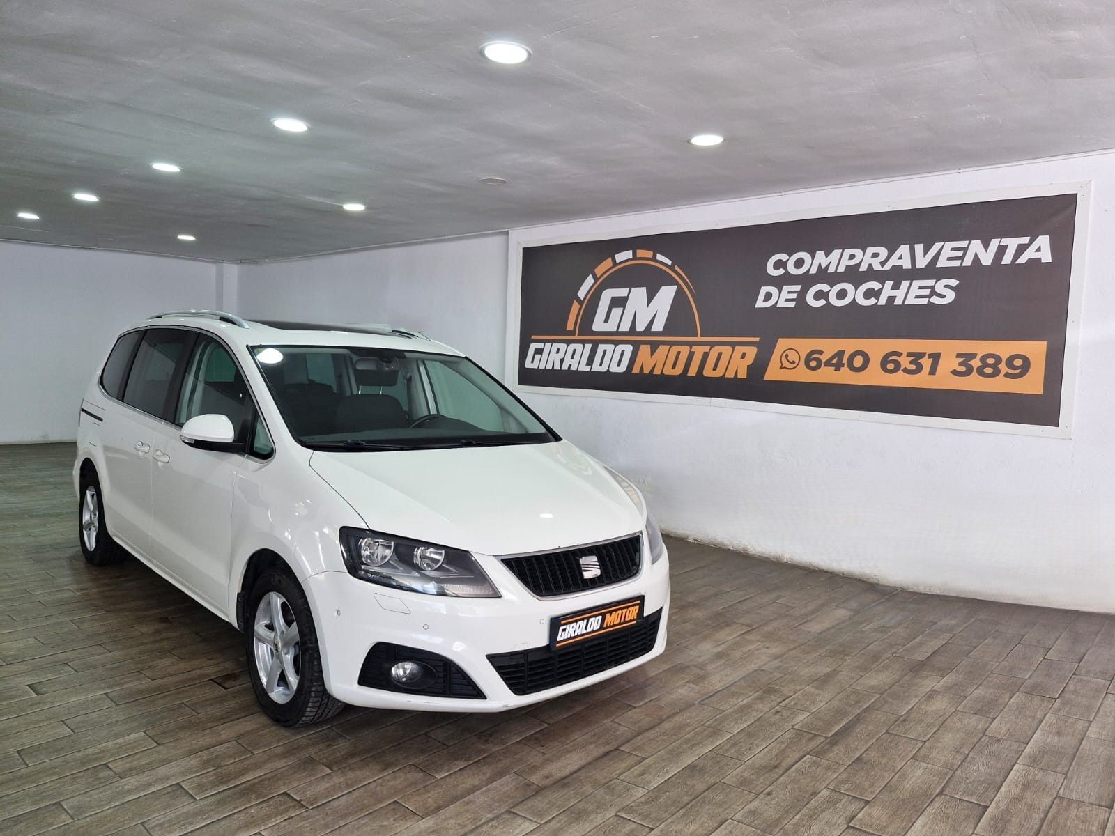 Minivan SEAT Alhambra de color blanco en la sala de exposición de un concesionario de automóviles, junto a un cartel de un concesionario de automóviles.