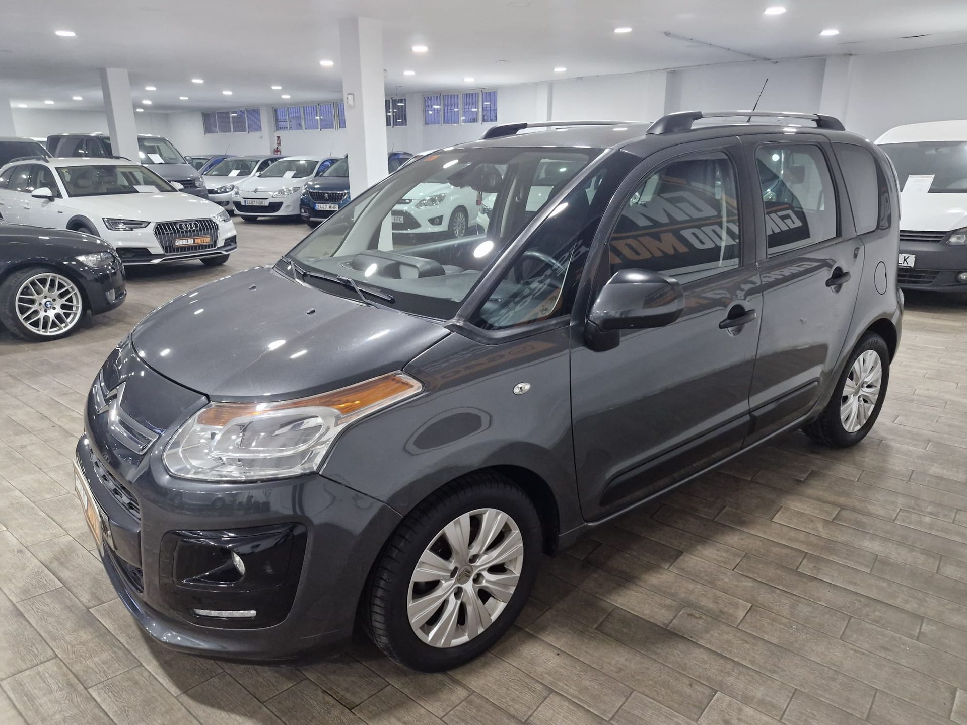 Citroën C3 Picasso gris en una sala de exposición, con otros coches visibles al fondo.