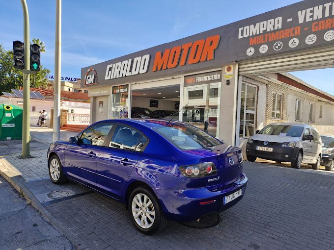 Mazda 3 azul estacionado frente al concesionario Giraldo Motor. Día soleado, calle, con coches al fondo.