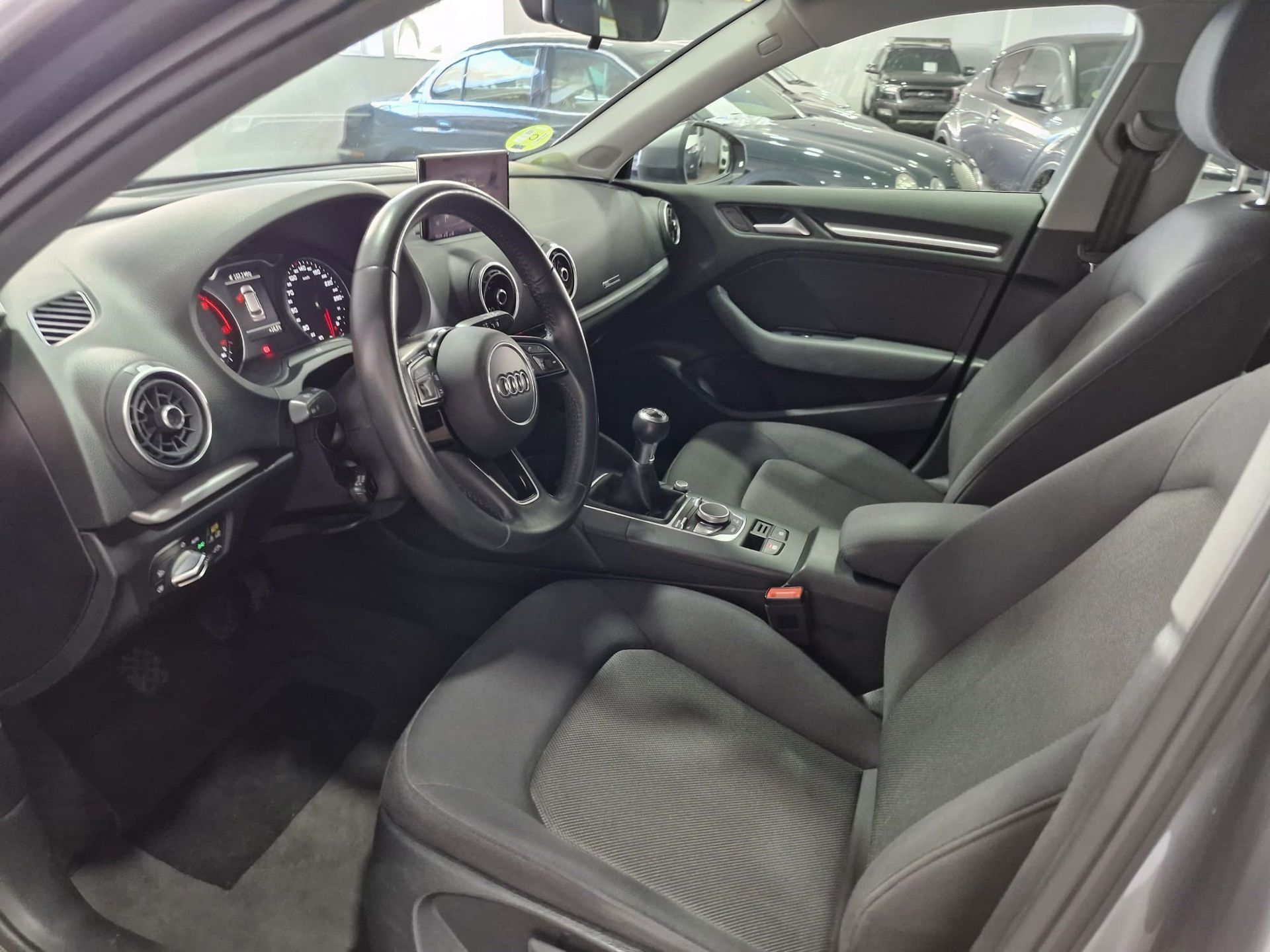 Interior de un Audi A3. Asientos, volante y salpicadero negros. Pantalla y palanca de cambios manual visibles.