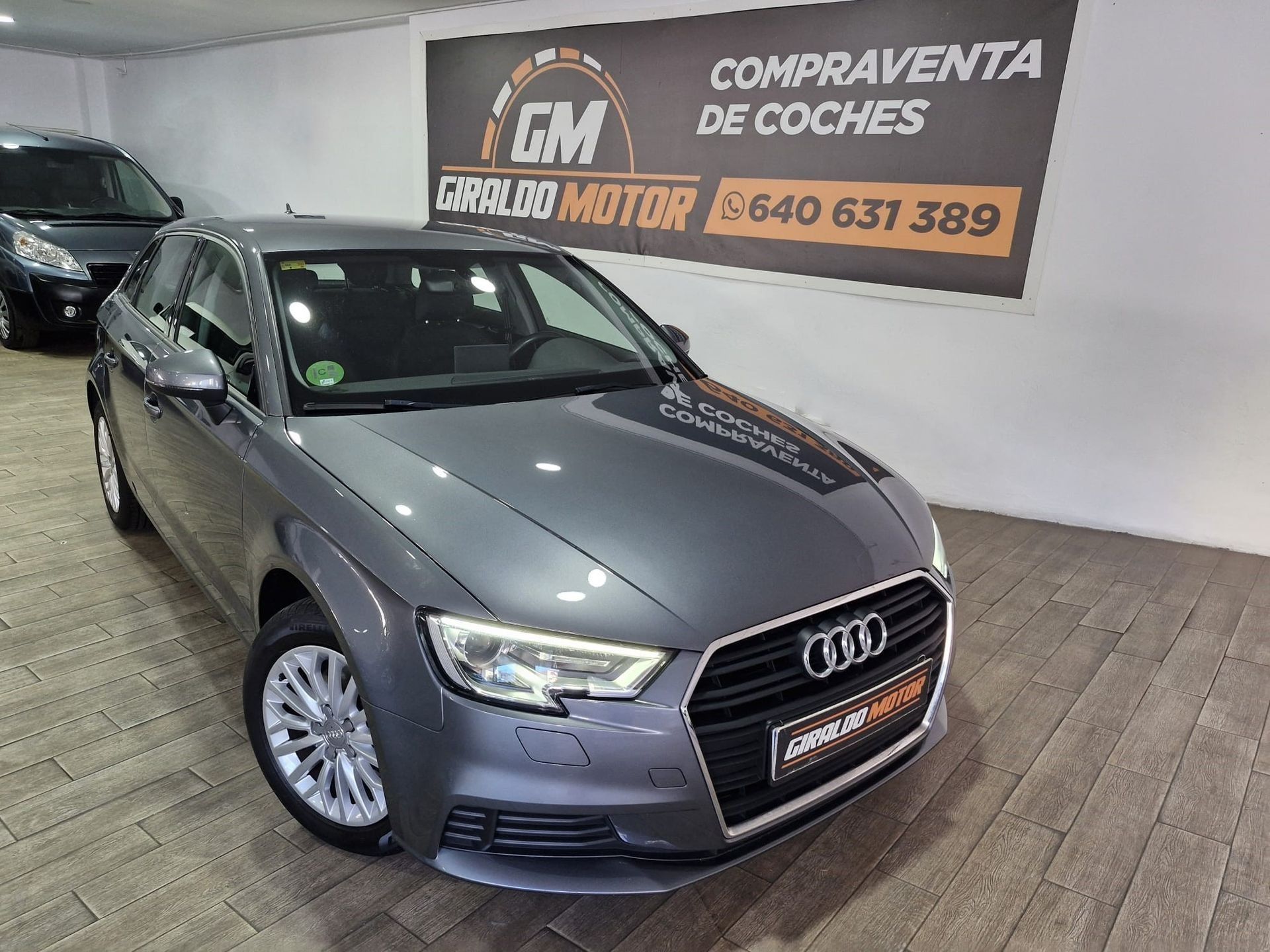 Un sedán Audi gris estacionado dentro de un concesionario de automóviles, con el logotipo del concesionario y el número de teléfono visibles.
