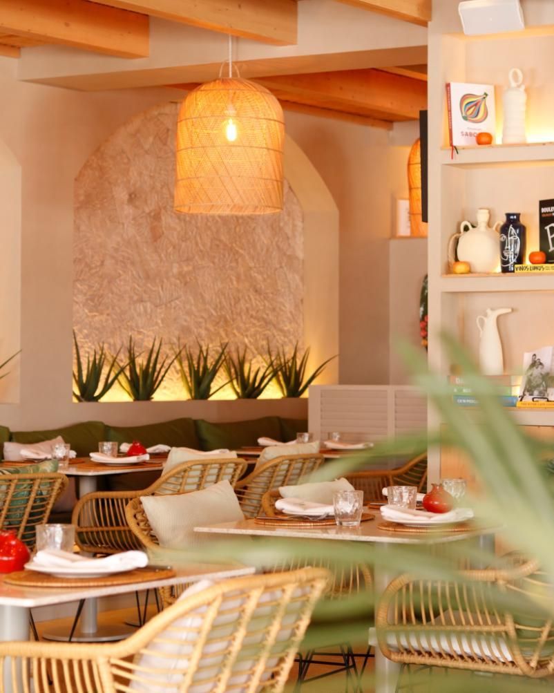 Interior del restaurante: sillas de ratán, mesas con platos blancos, lámpara colgante tejida, pared texturizada