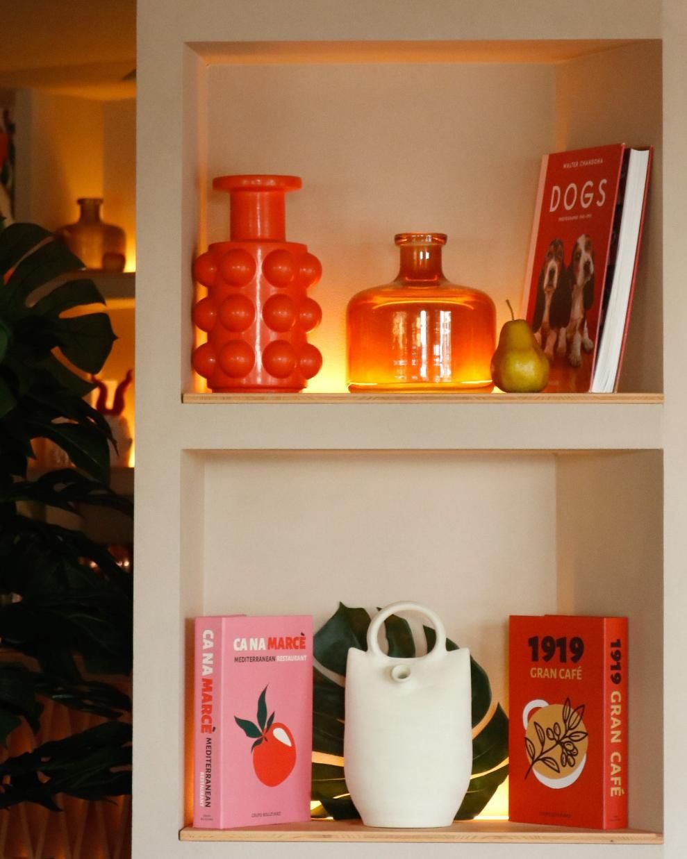 Estanterías con jarrones naranjas y blancos, un libro y una pera; iluminados desde atrás.