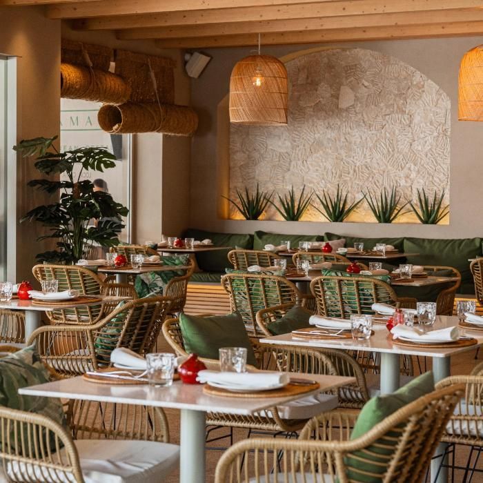 Interior de restaurante con sillas de mimbre, mesas cuadradas y lámparas tejidas.