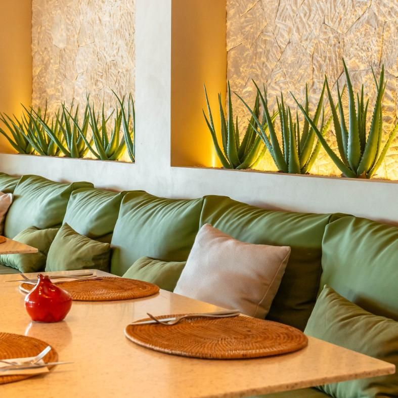 Cabina de restaurante con cojines verdes, plantas de aloe en pared texturizada y puestas de mesa.