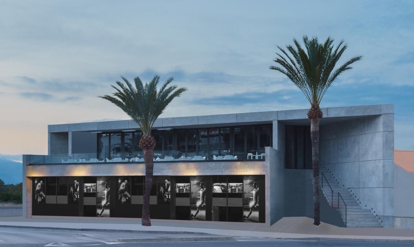 Edificio moderno de hormigón con palmeras en el frente, ventanas en la planta baja, terraza arriba.