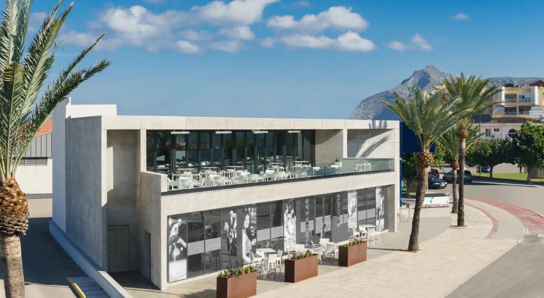 Edificio moderno de dos pisos con balcón de vidrio, palmeras y fondo de montaña en un día soleado.