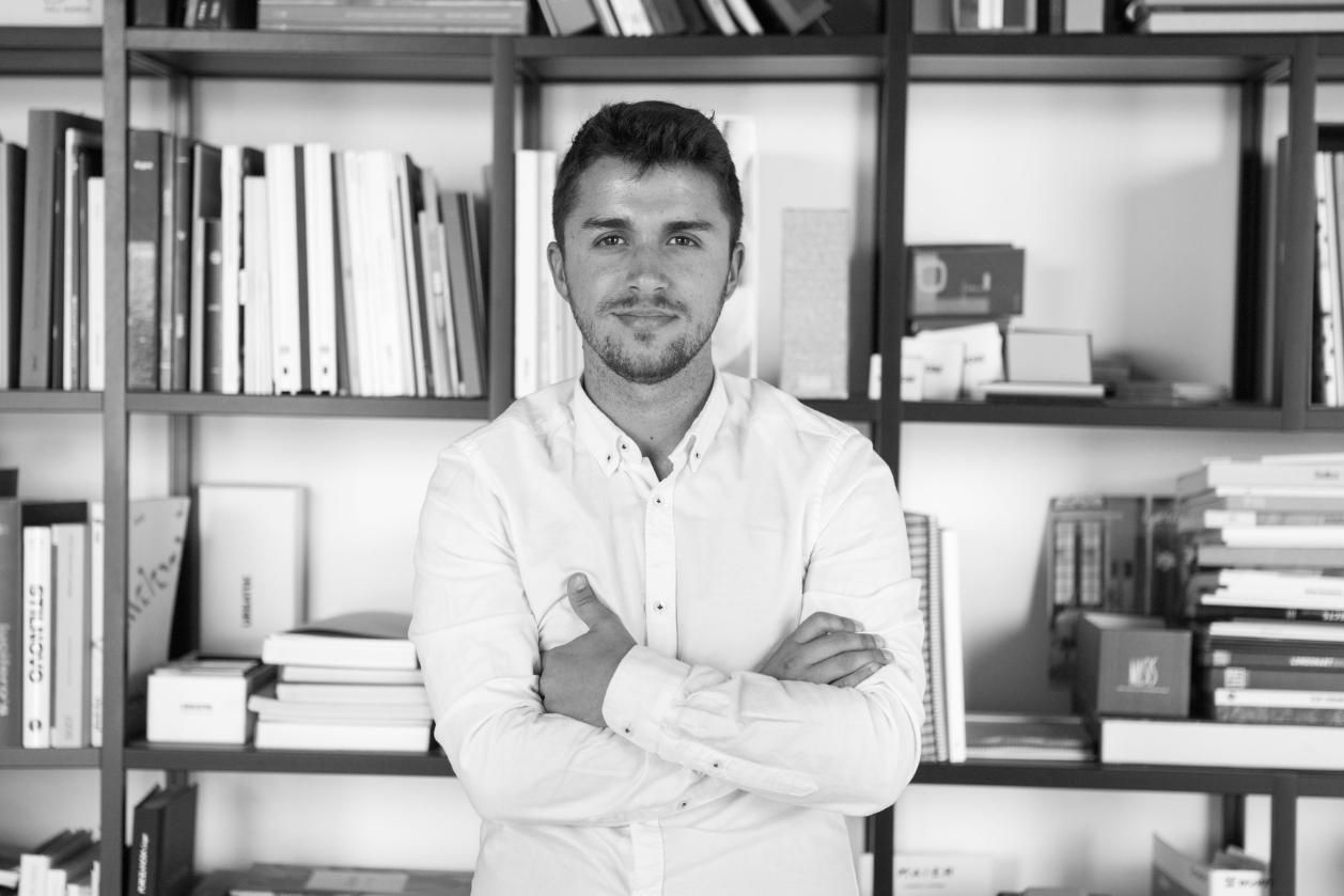 Hombre con los brazos cruzados, delante de una estantería llena de libros y objetos.