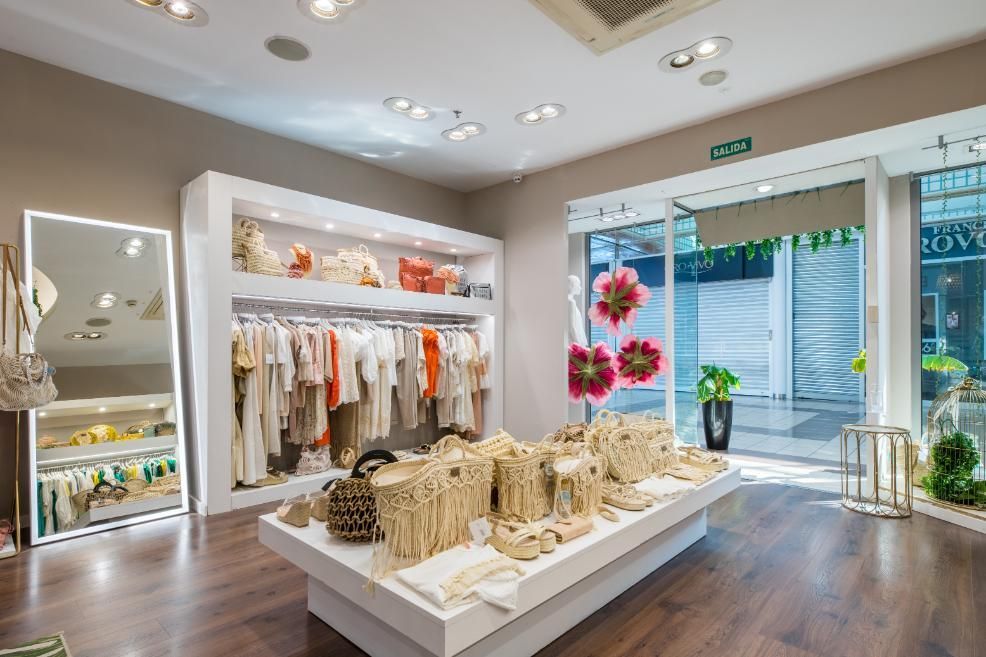 Interior de una tienda de ropa con ropa en exhibición, un gran espejo y puertas abiertas al exterior.