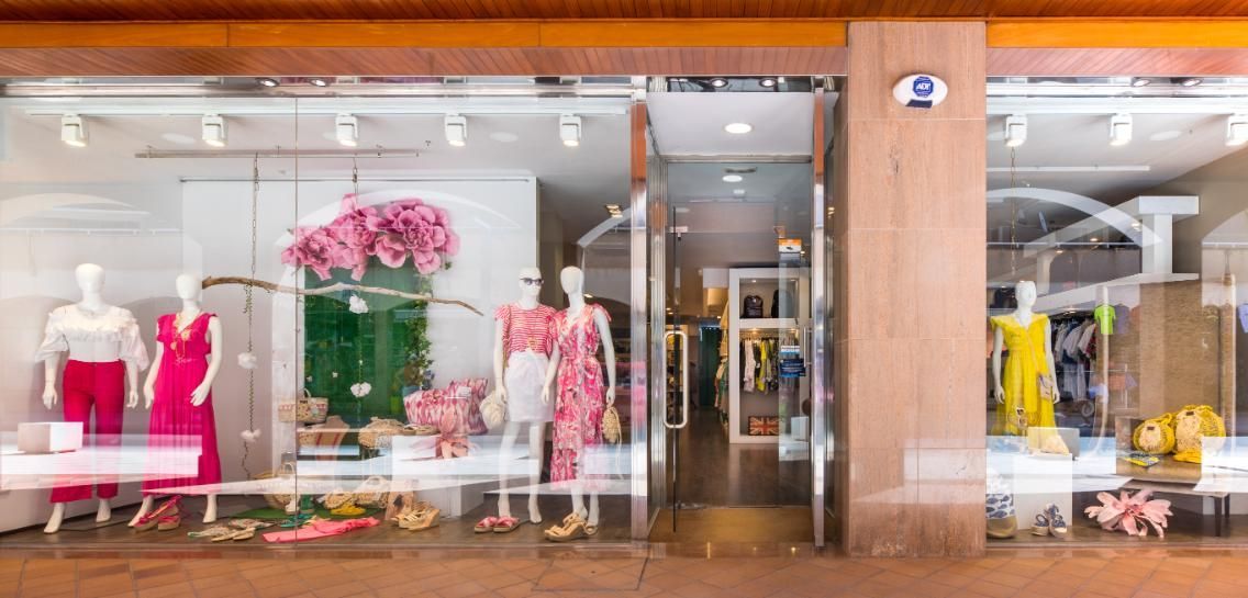 Fachada de tienda con maniquíes exhibiendo ropa. Trajes rosas y blancos, arreglos florales. Puerta de cristal.