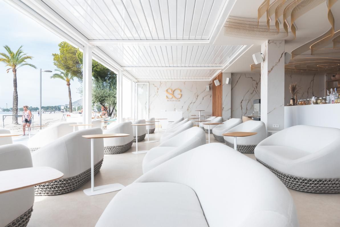 Sala de estar blanca con vista a la costa, con asientos curvos, mesas pequeñas y un techo de listones.