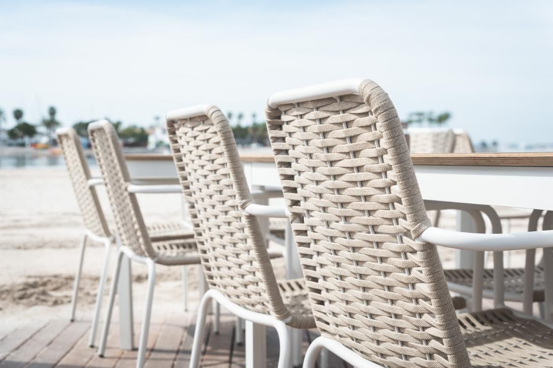 Sillas con respaldo tejido se encuentran alrededor de una mesa blanca en una terraza junto a la playa.