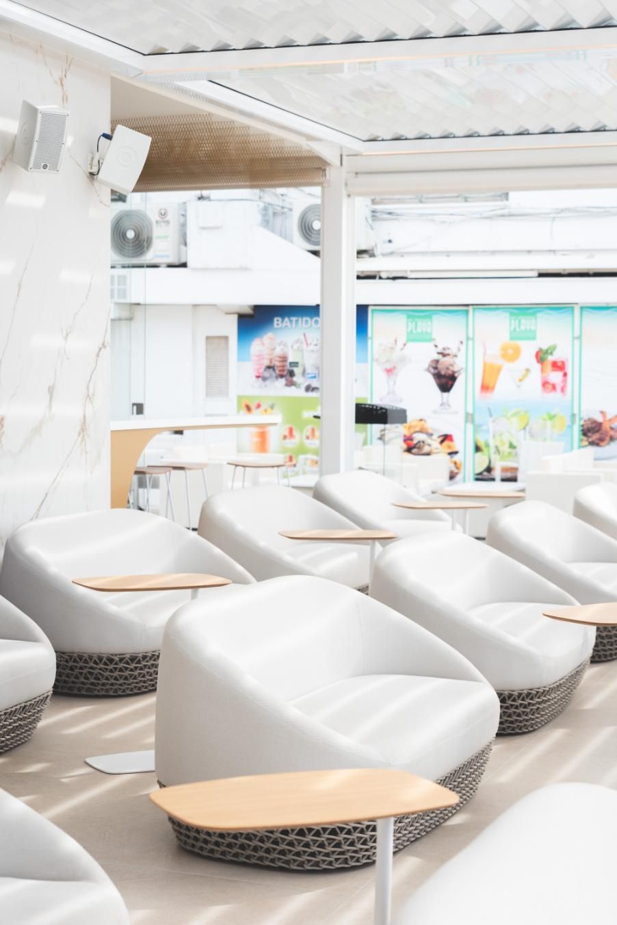 Sillones blancos con mesas pequeñas bajo un techo de listones en un café con carteles de comida al fondo.