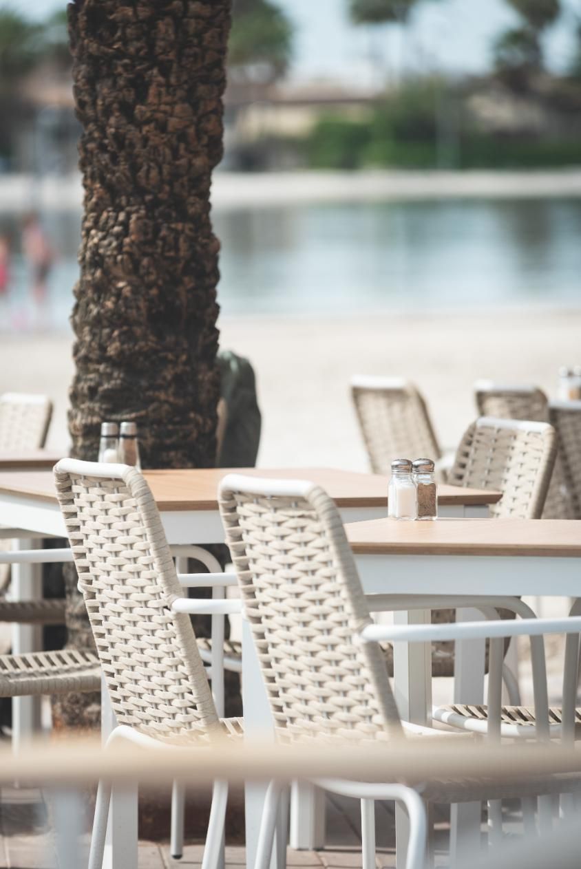 Cafetería junto a la playa con mesas y sillas blancas, tronco de palmera texturizado y vista borrosa del océano.