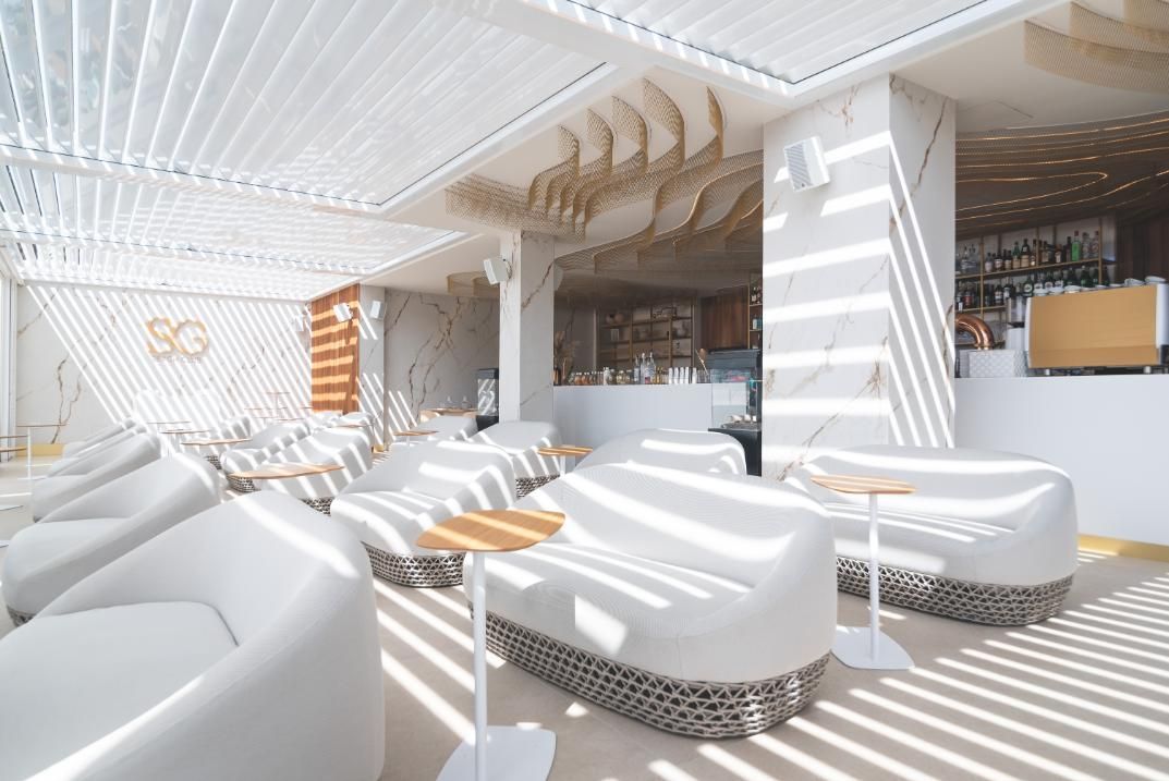 Zona de estar moderna y luminosa con sofás blancos curvos, mesas pequeñas y un bar al fondo.