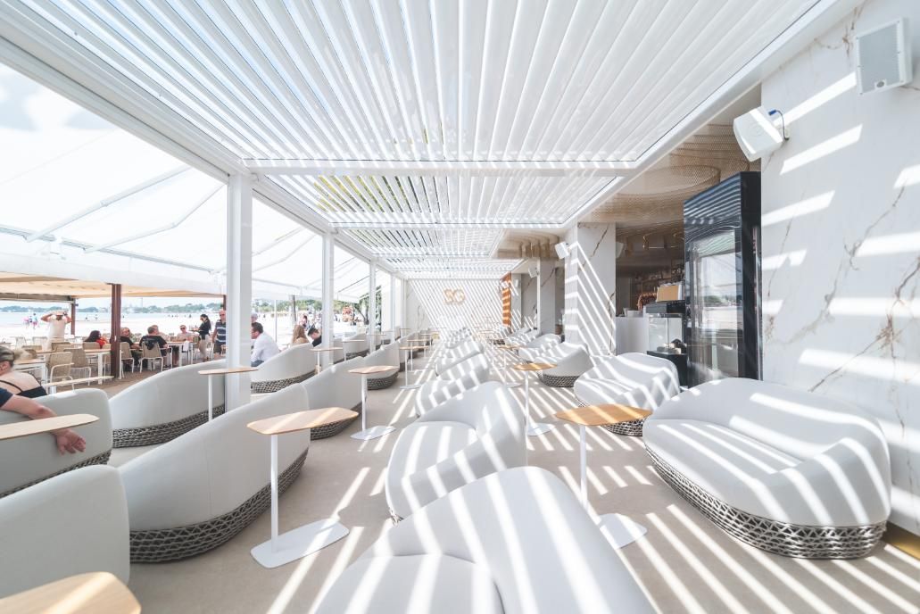 Zona de estar al aire libre de color blanco con techo de listones; el sol proyecta sombras rayadas.