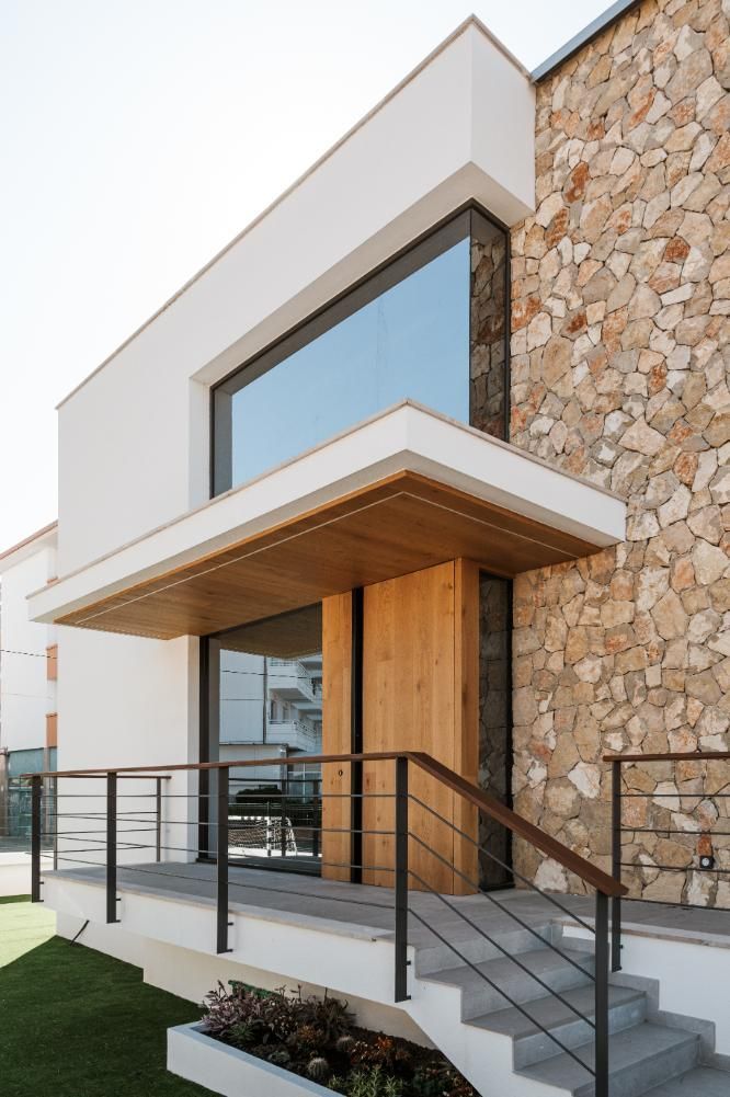 Exterior de casa moderna con fachada blanca, pared de piedra, puerta de madera y ventana grande.
