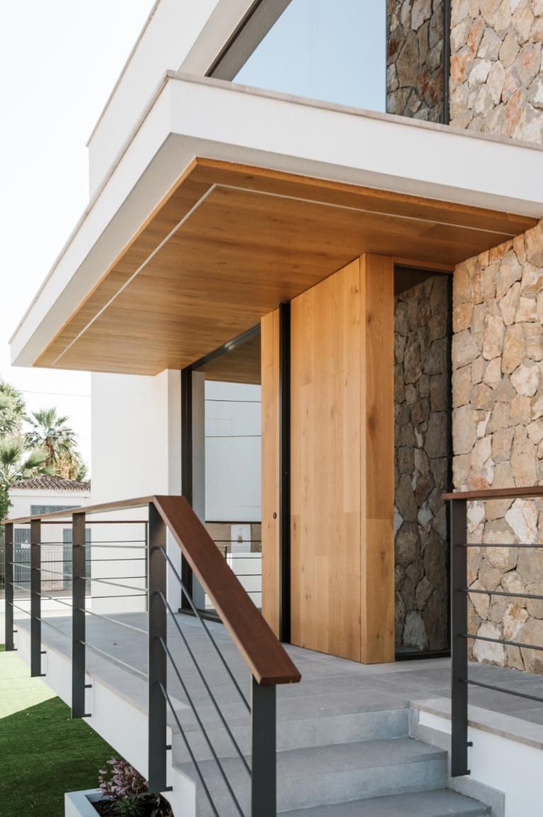 Entrada de casa moderna con puerta de madera, pared de piedra y barandillas de metal.