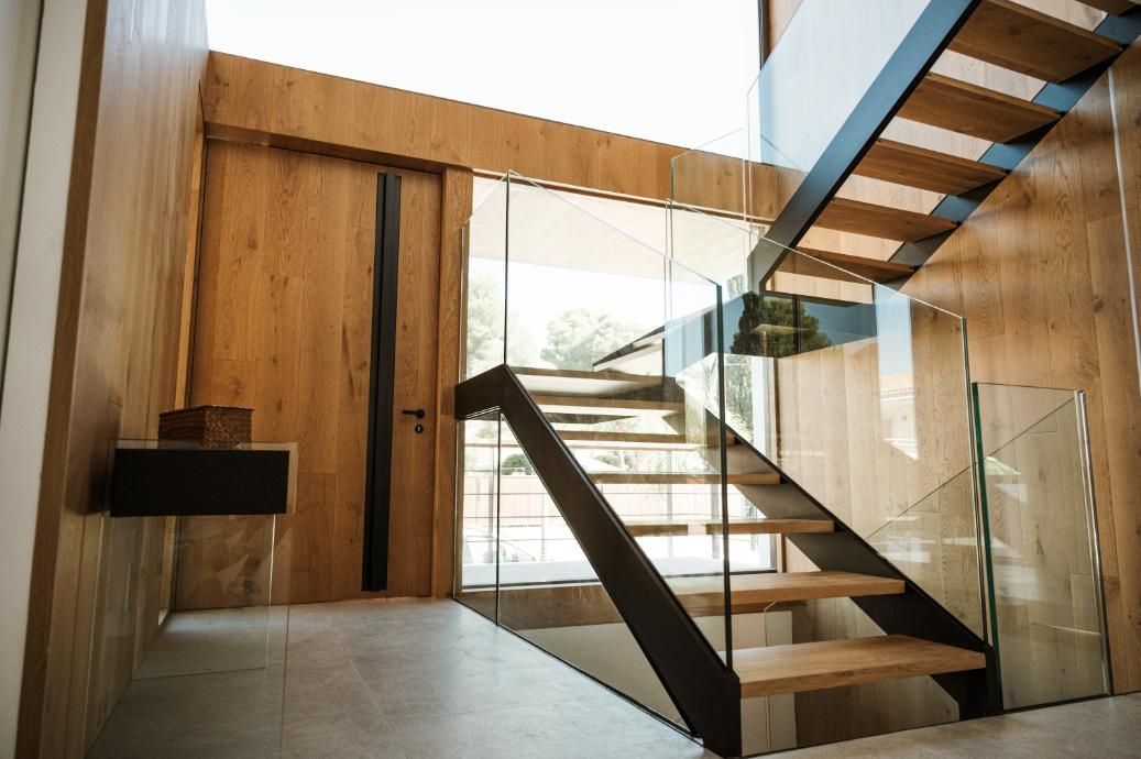 Escalera de madera con barandilla de cristal en una entrada moderna y luminosa con paredes de paneles de madera.