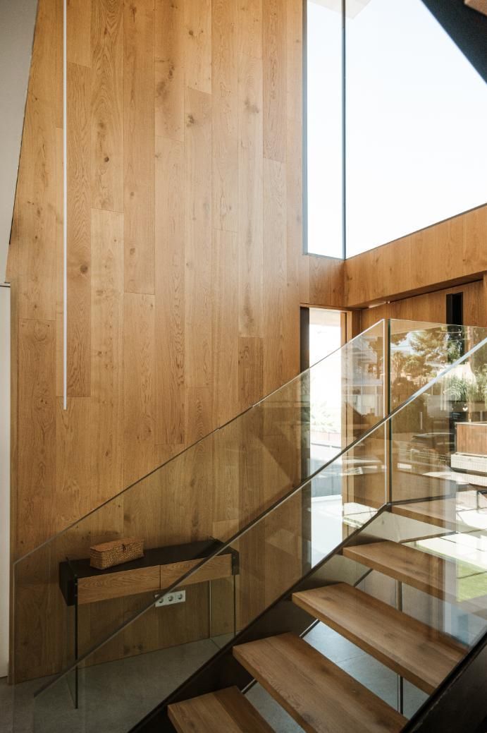 Escalera de madera con barandilla de cristal; luz natural de un gran ventanal ilumina el espacio.