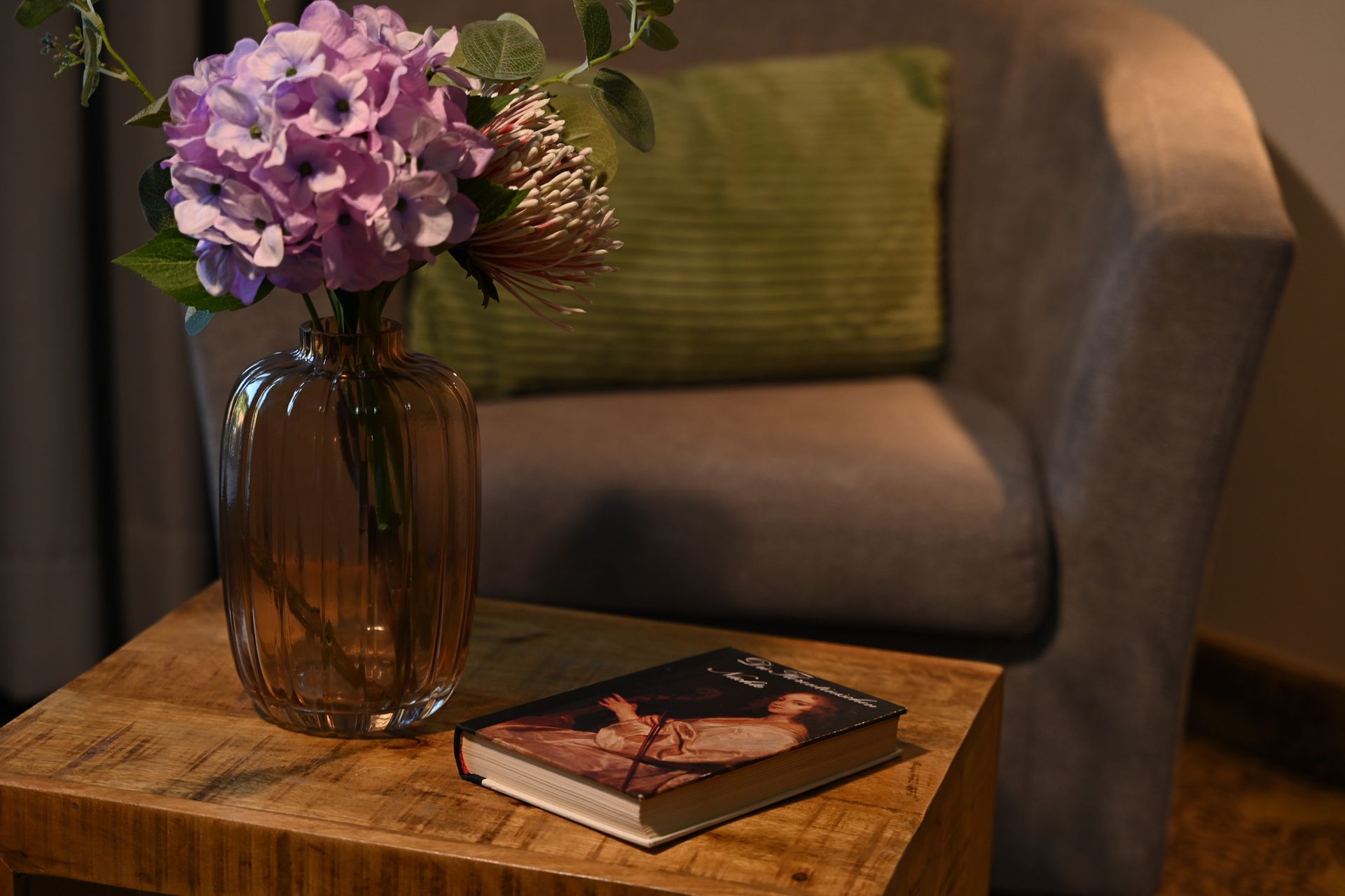 Eine Vase mit lila Blumen, ein Buch und ein grünes Kissen auf einem hölzernen Beistelltisch neben einem Sessel.