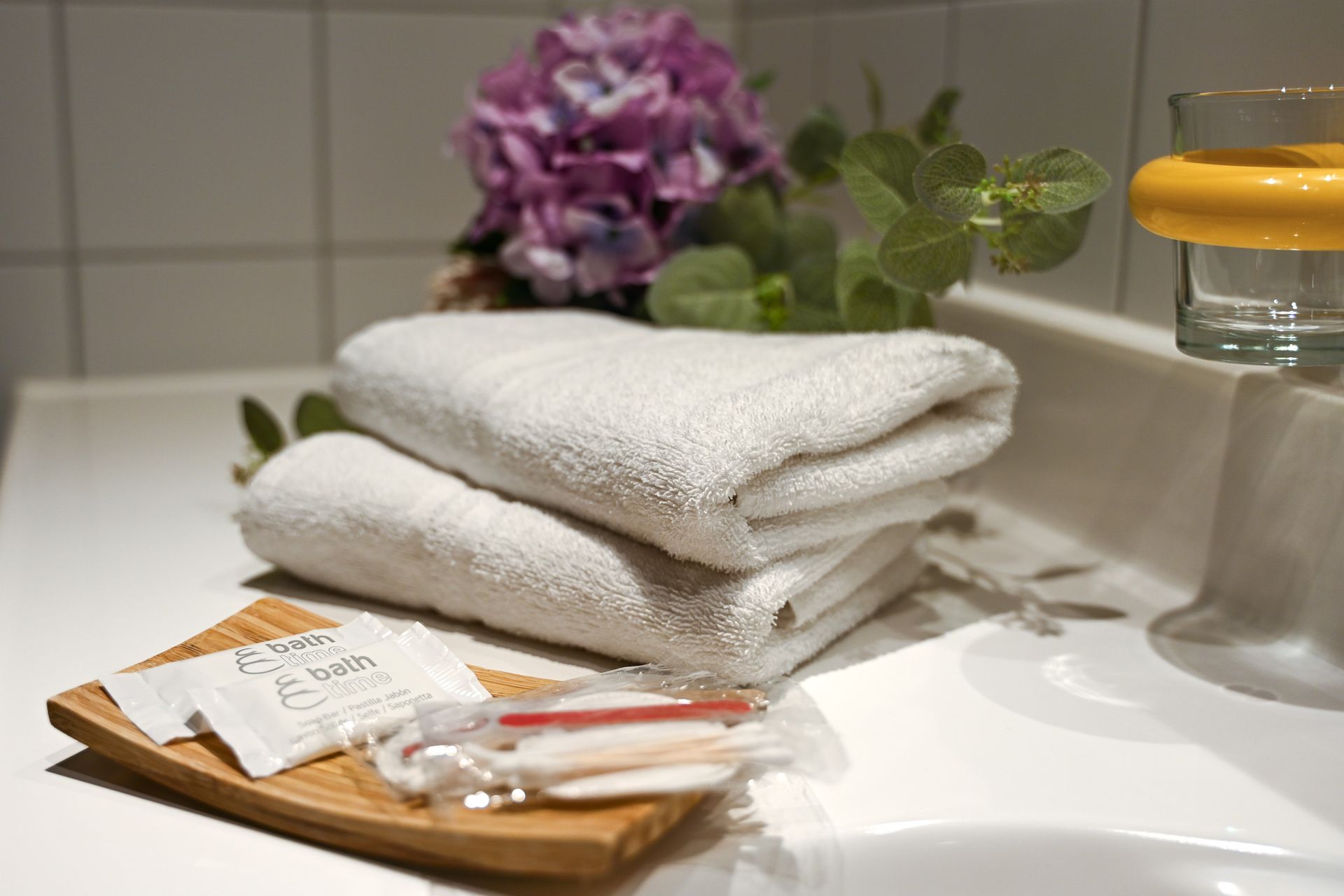 Weiße Handtücher lagen auf einem Waschtisch im Badezimmer, dazu Toilettenartikel und ein lila Blumenarrangement.