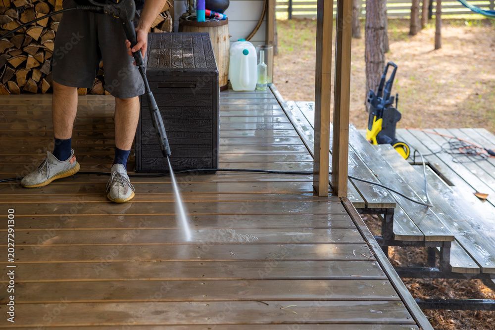 Eine Person reinigt eine hölzerne Veranda neben einem Haus mit einem Hochdruckreiniger, wobei das Wasser über die Veranda gesprüht wird.