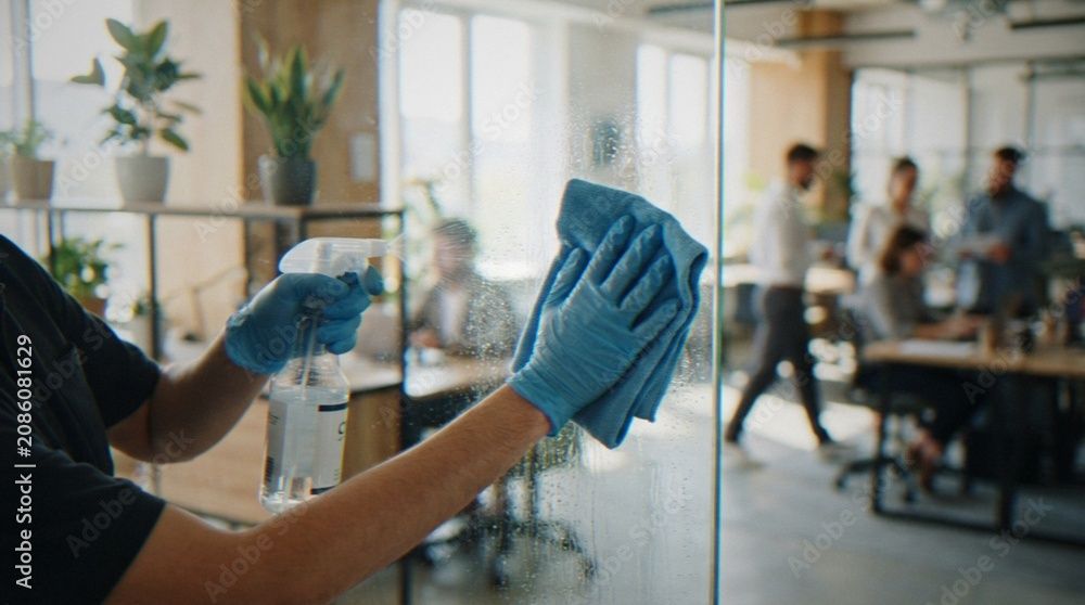 Eine Person mit blauen Handschuhen wischt eine Glastrennwand in einem geschäftigen Büro ab.