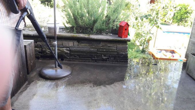 Eine Person reinigt eine hölzerne Veranda neben einem Haus mit einem Hochdruckreiniger, wobei das Wasser über die Veranda gesprüht wird.