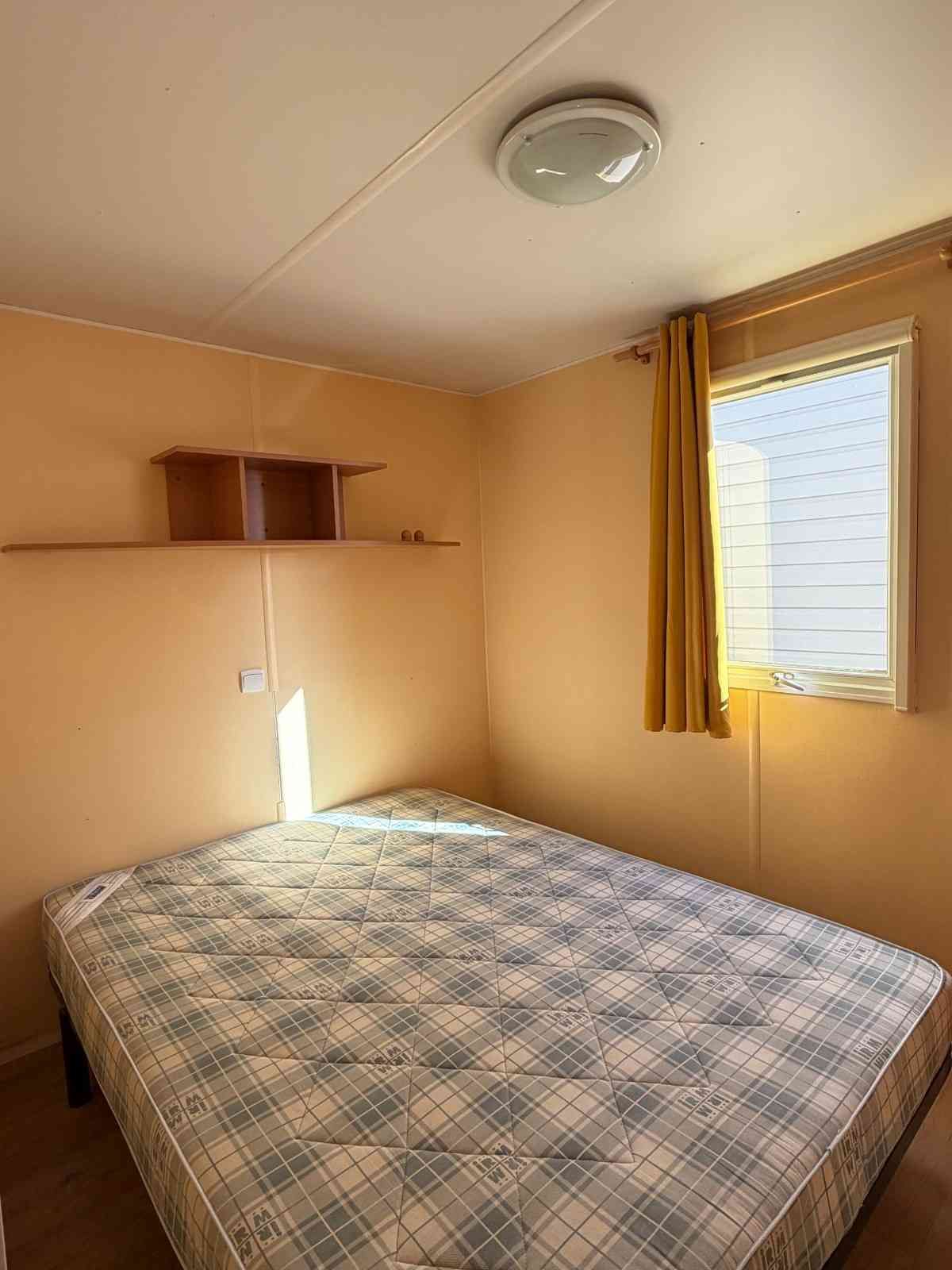 Habitación pequeña con cama, estante de pared, lámpara de techo y ventana con cortinas amarillas.