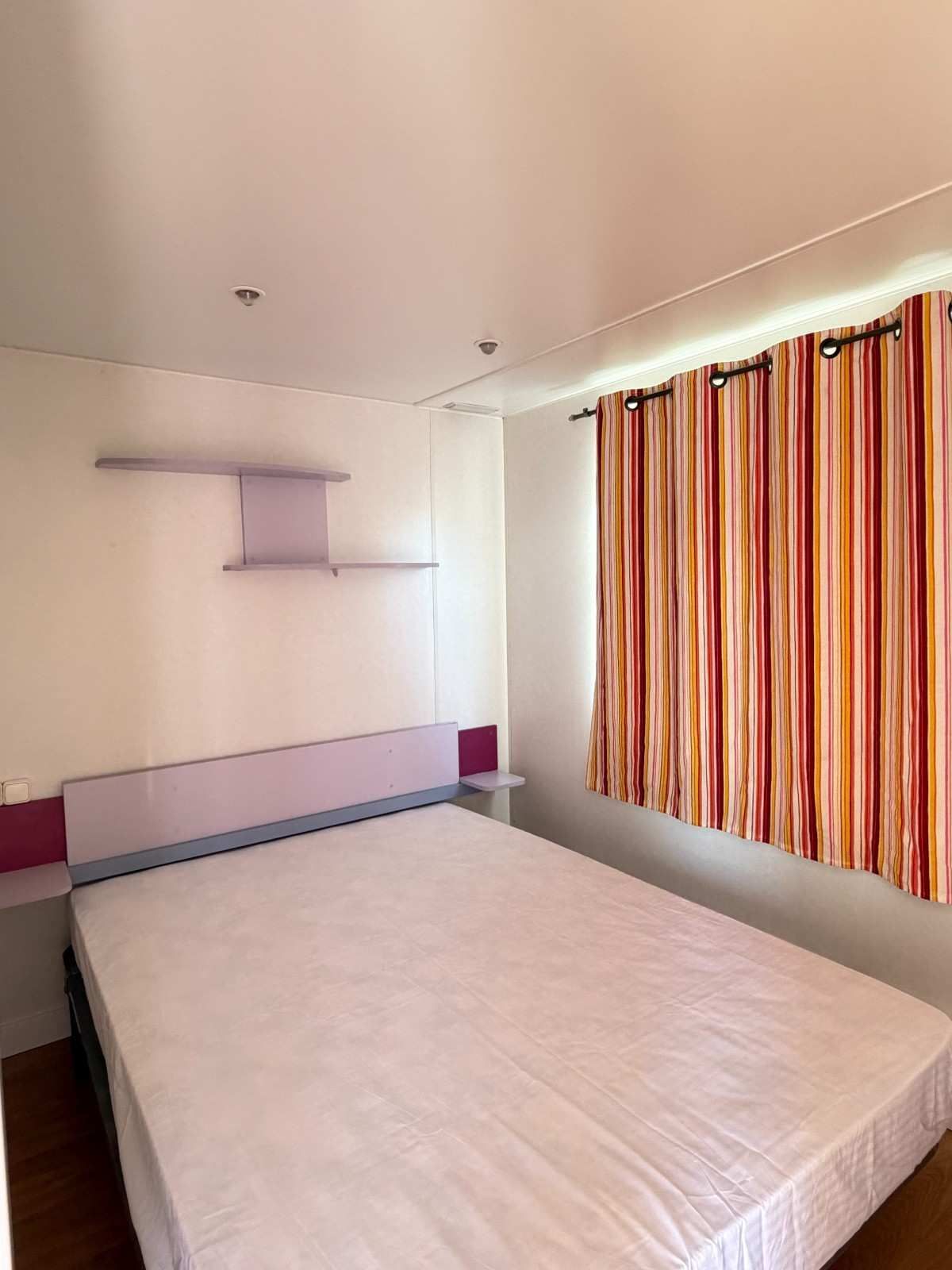 Habitación pequeña con cama, estantes en la pared y cortinas a rayas que dejan pasar la luz natural.