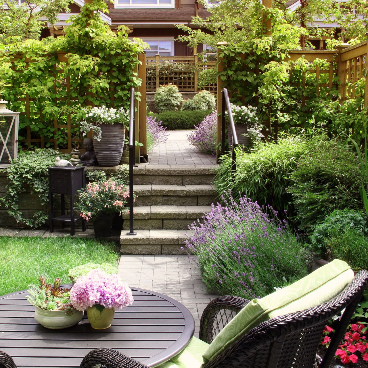 Patio de jardin luxuriant avec marches en pierre, fleurs en pot et sièges en osier autour d'une table ronde.