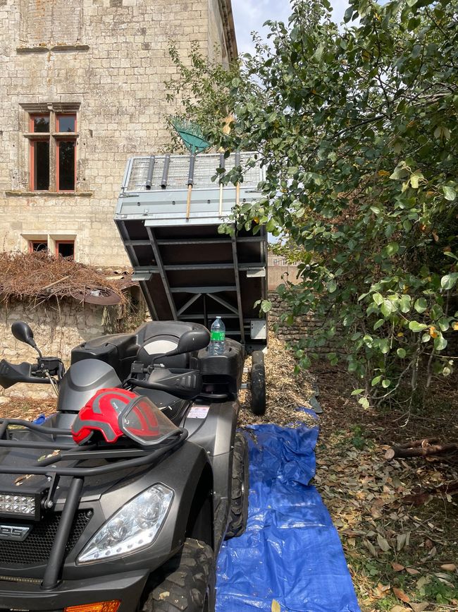 Tracteur noir garé près d'un muret en pierre et d'un abri, partiellement recouvert d'une bâche bleue sous des arbres