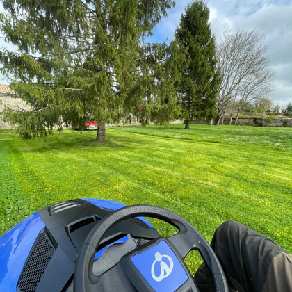 Une personne conduit une tondeuse bleue dans un parc verdoyant et ensoleillé, avec des arbres et de l'herbe.